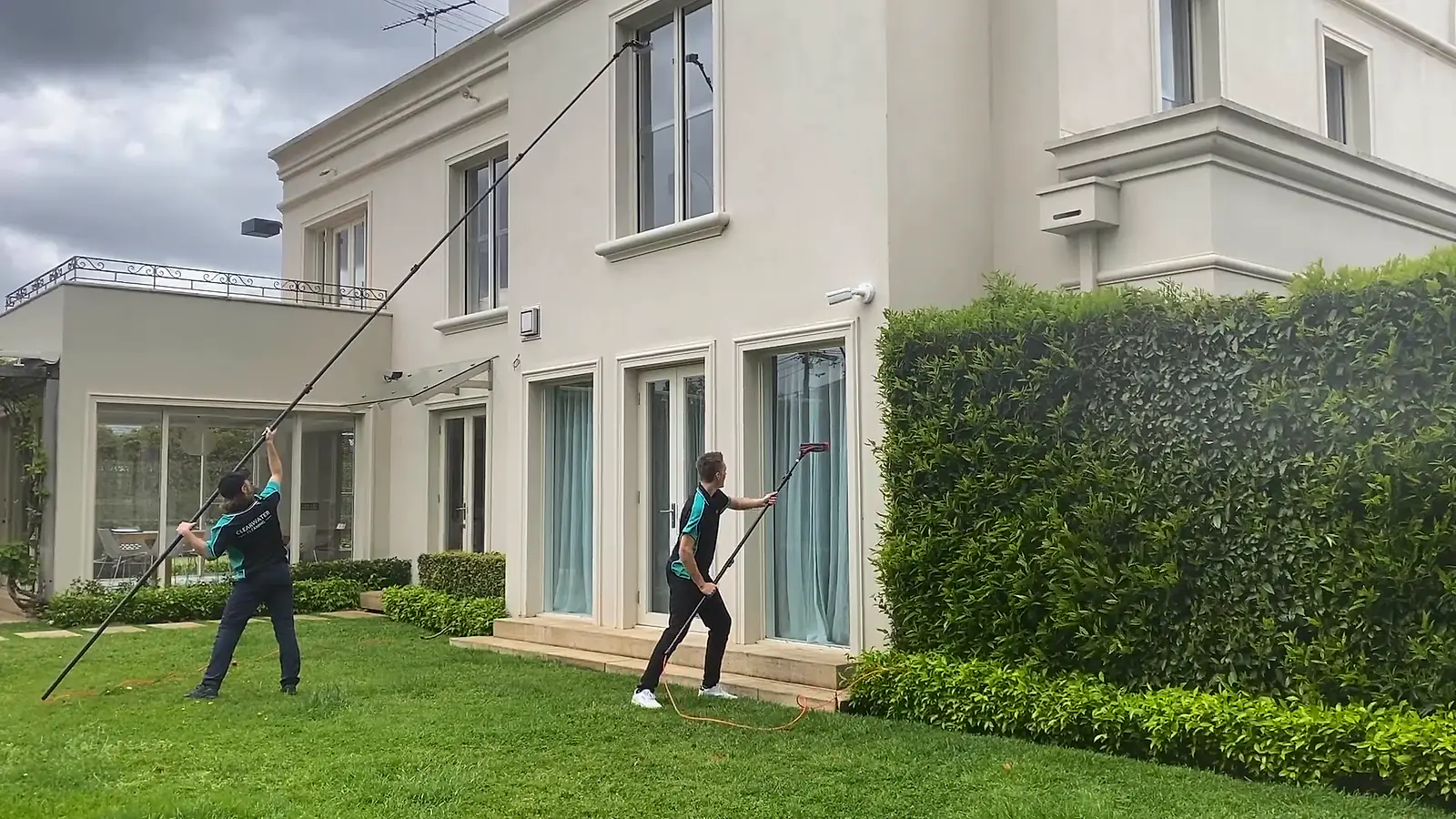 Two Clearwater cleaners reaching second-storey windows from the ground with water-fed poles