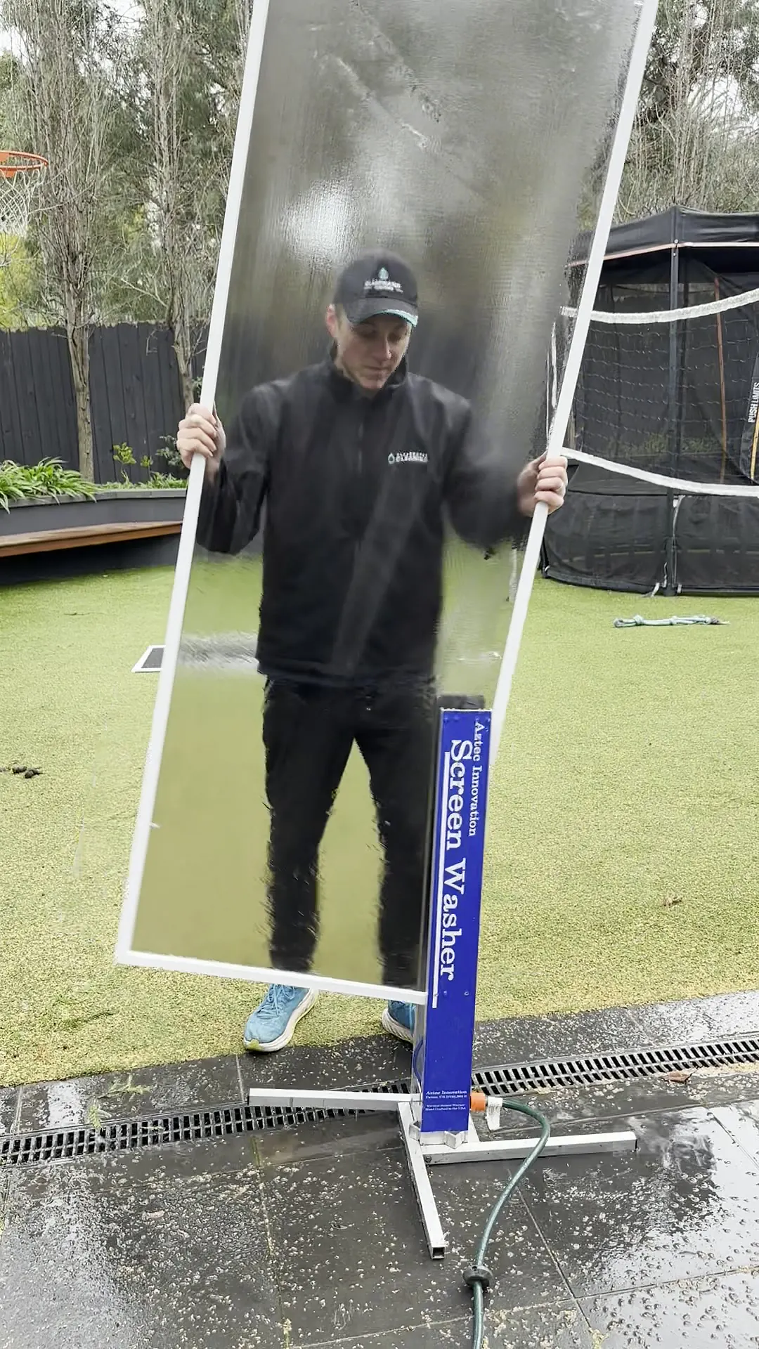 Fly screen lifted from a screen-washing machine with water sheeting off it, the frame held by a Clearwater cleaner