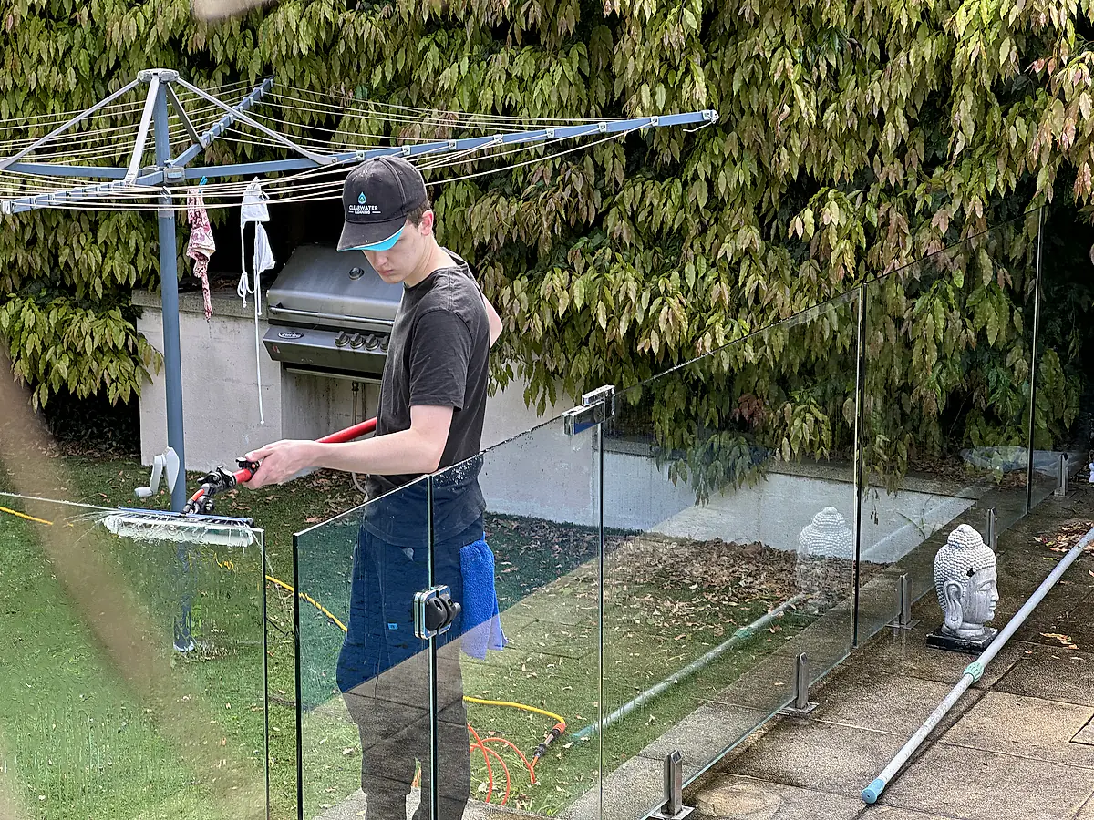 Clearwater cleaner running a water-fed pole along a line of glass pool fence panels in a back garden